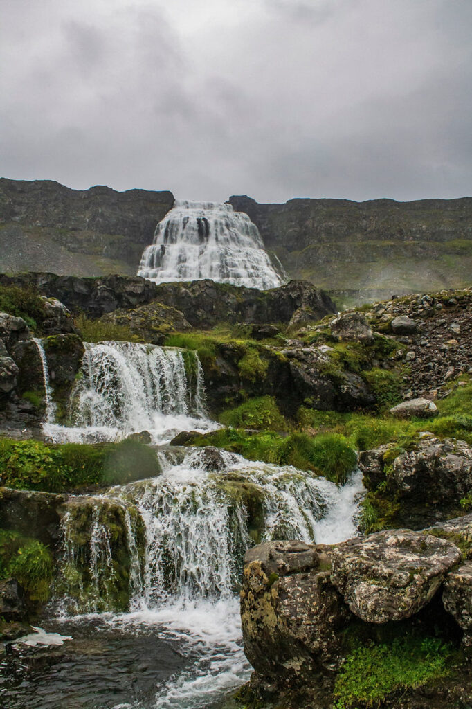 secret iceland elopement spots: Waterfall cascading down a rocky, mossy landscape under a cloudy sky. The scene is lush and serene, with vibrant greens and greys.