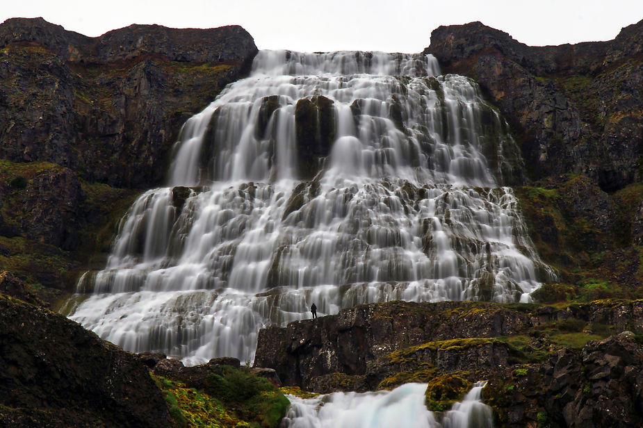 Tall waterfall cascading over rocky cliffs; a lone person stands at the base, emphasizing scale. Lush greenery surrounds the scene.