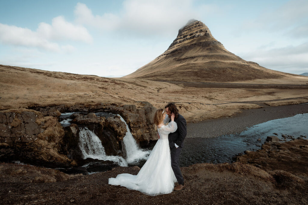 iconic iceland wedding location: A couple embraces by a waterfall, with a mountain in the background. The bride wears a white elopement dress.