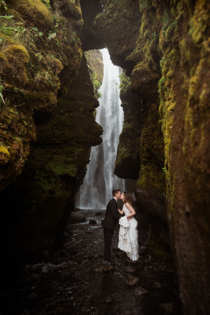 secret iceland elopement spots: bride and groom kiss near a cascading waterfall, framed by moss-covered cliffs, creating a romantic and serene scene.