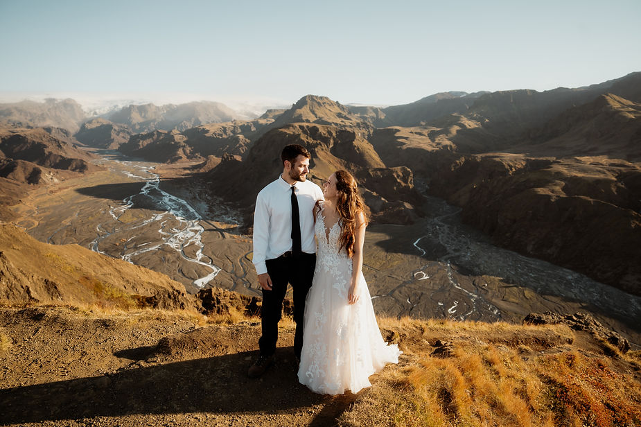 secret iceland elopement spots: groom and bride smiling at each other on a mountain with a river below. Clear sky and rugged terrain in the background.