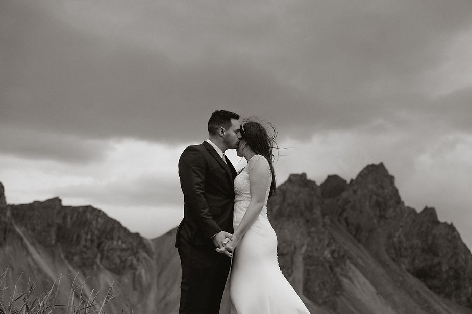 bride and groom in front of rocky iceland mountains. The woman wears a white dress, the man a dark suit. The mood is romantic and serene.