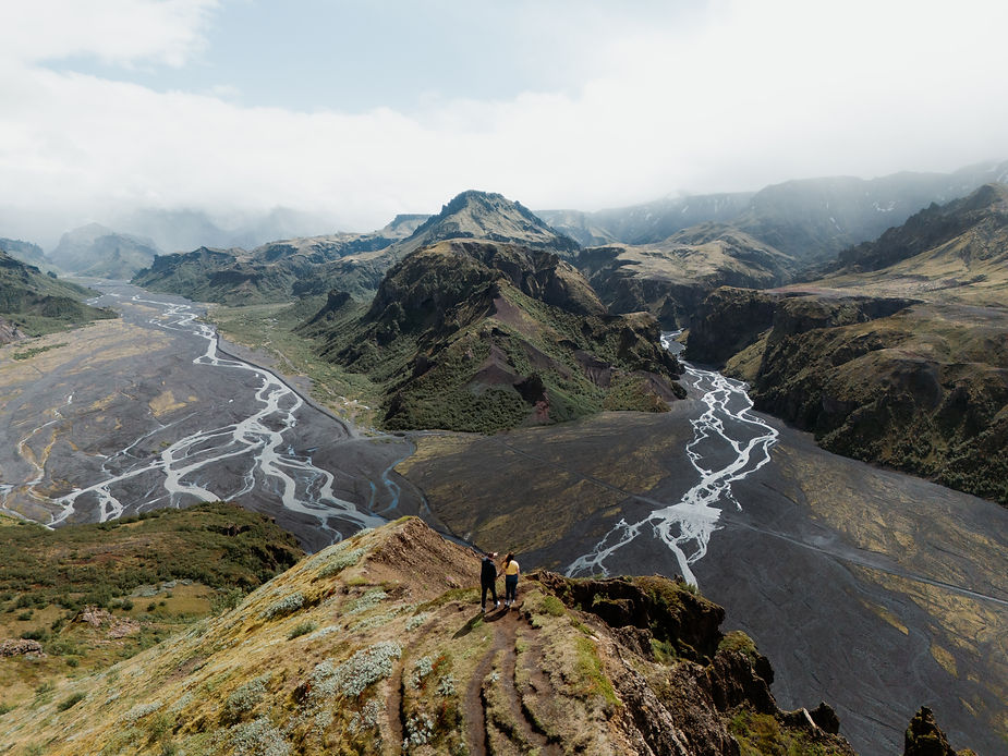 Two people stand on a grassy cliff overlooking a vast river canyon and distant mountains under cloudy skies, evoking a sense of adventure in iceland