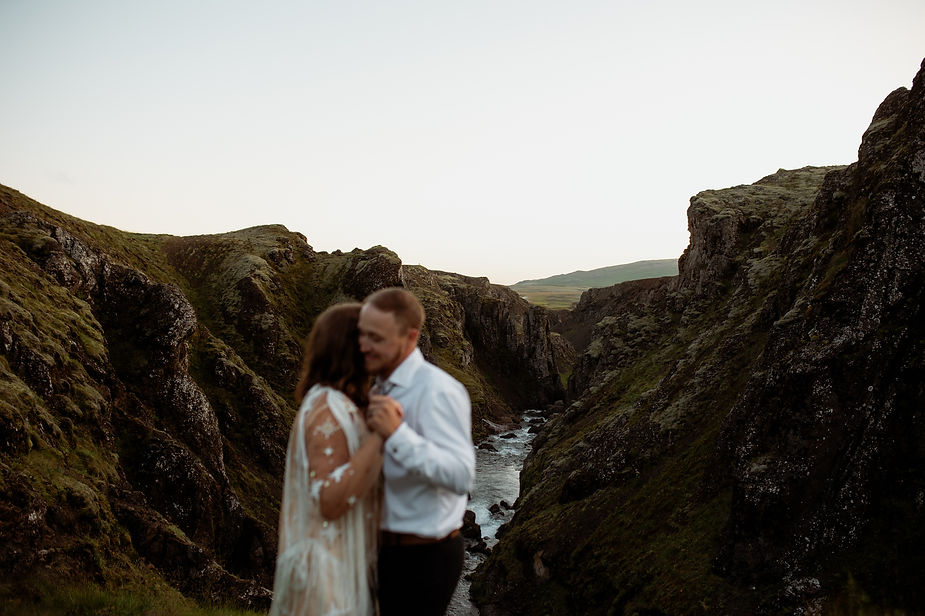 secret iceland elopement spots: bride and groom embraces at a cliff's edge, with a river in the valley below. The sky is clear, and the landscape is lush and rugged, evoking romance.
