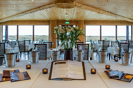 iceland wedding venue, Elegant restaurant with wood ceiling and large windows overlooking a field. Tables with white cloths, flowers, menus, and lit candles.