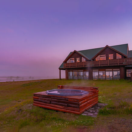 iceland wedding venue with a green roof at sunset, steaming wooden hot tub in foreground, surrounded by grassy landscape under a purple-pink sky.