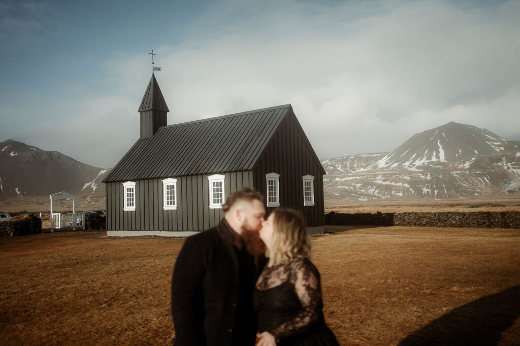 iconic iceland wedding location: A couple kisses in front of a black church with white windows. Snowy mountains in the background. Overcast sky and dry grass.