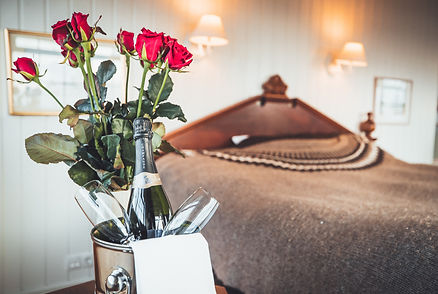 Red roses in a vase and champagne with glasses on a table in a cozy bedroom at iceland wedding venue. Warm lighting, brown bedspread, and wall lamps visible.