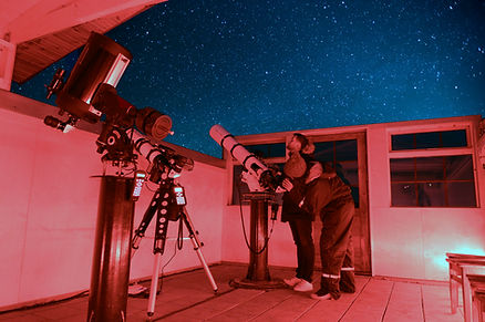 Two people stargazing with telescopes in a red-lit observatory, under a clear, starry sky. The mood is focused and serene.