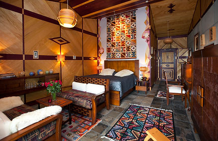 Cozy room with geometric wooden walls, colorful patterned rugs, a bed with pillows, and chairs with cushions. Roses on a table add warmth.