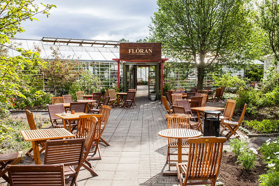 Outdoor iceland wedding venue with wooden tables and chairs on a patio, surrounded by lush greenery and a greenhouse. Sign reads "Floran Cafe Bistro." Sunny and serene.
