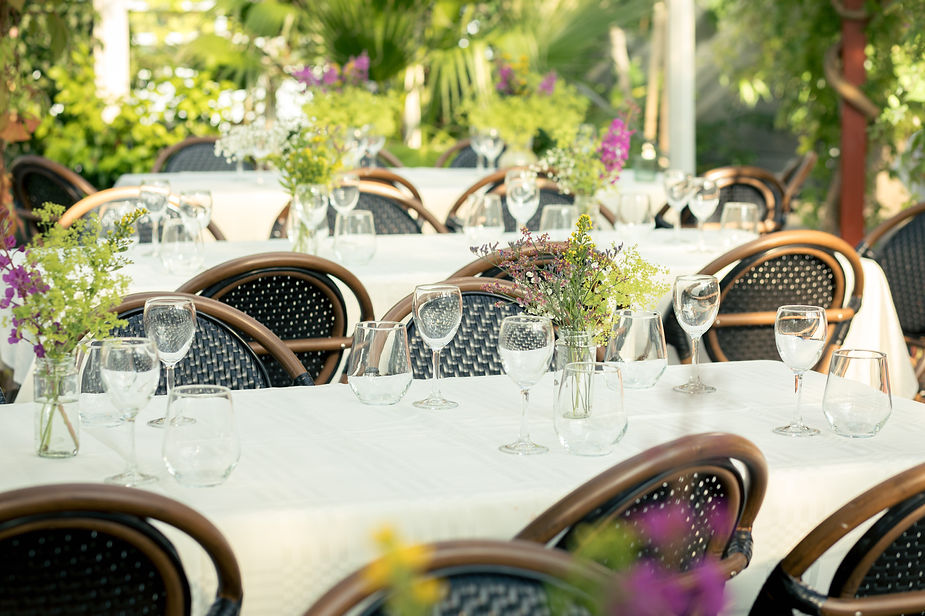 Outdoor wedding reception area with white tablecloths, empty wine glasses, and vases of wildflowers on tables. Wicker chairs, lush greenery.