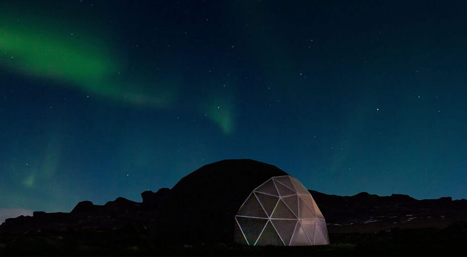 Geodesic dome under vibrant green Northern Lights in a clear starry night sky, set against a dark mountainous silhouette. iceland wedding venue