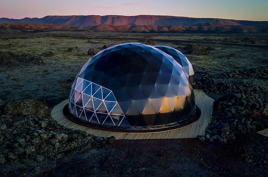 Geodesic dome structure on rocky terrain at sunset, with mountains in the background. The dome has a geometric pattern and reflects warm hues and is an iceland wedding venue.