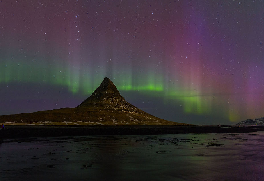 Northern lights in green and purple illuminate the night sky over a pointed mountain, reflecting in the dark water below.