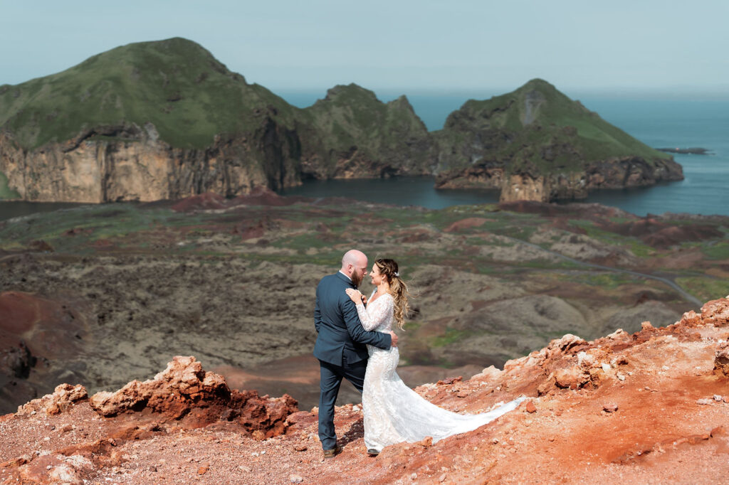 iconic iceland wedding location: A couple in wedding attire embraces on a reddish rocky cliff with green mountains and blue ocean in the background.