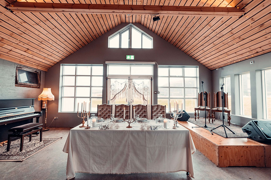 Elegant wedding venue dining setup with a white tablecloth, candles, and tufted chairs. A cozy wooden ceiling, piano, and empty stage create a warm ambiance.