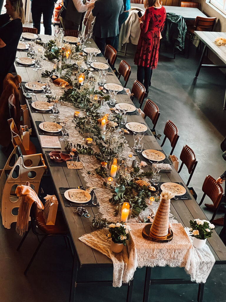 Elegant wedding reception table setting with plates, glasses, and candles. Greenery and flowers adorn the table. People stand and chat in the background.