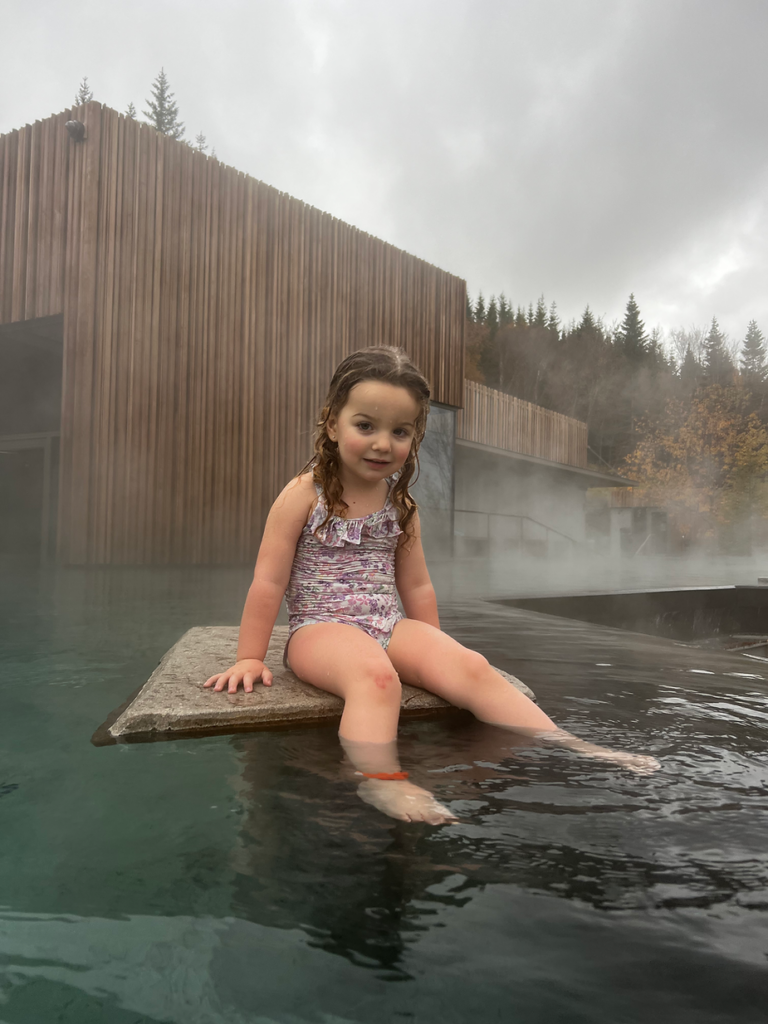 Young girl in a floral swimsuit sits at the edge of a steaming outdoor pool. Wooden building and trees are visible under a cloudy sky in iceland