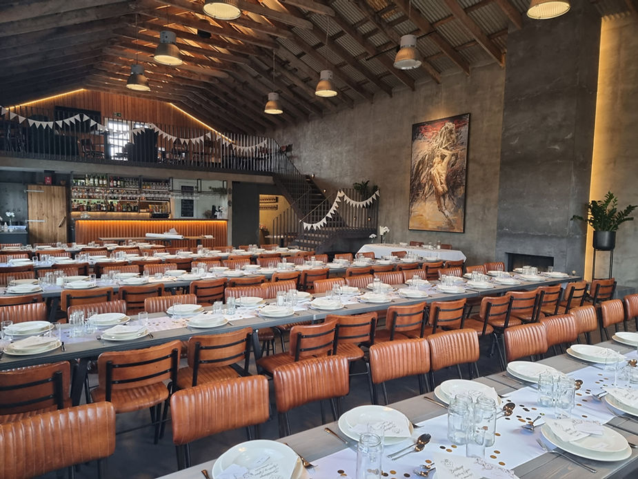 Rustic reception dining hall with long tables set for a banquet. Leather chairs, white plates, glasses, and cutlery. Large painting and bar in back.