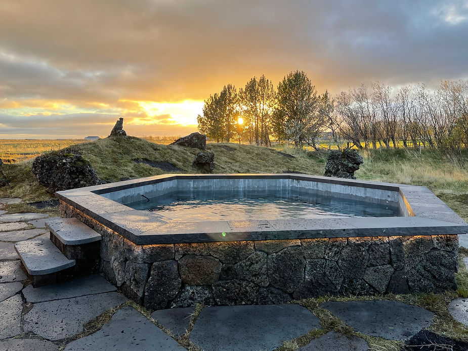 Stone hot tub in a grassy landscape during sunset, with trees and rocks in the background, creating a serene and warm atmosphere in iceland.