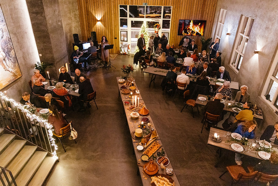 A wedding reception in a warmly lit room with guests seated at tables. A singer performs; a long table holds a variety of food.