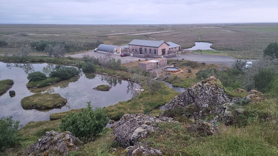 Rural landscape with a pond, lush greenery, and rocky terrain. Buildings, a car, and a tub are visible. Overcast sky creates a serene vibe at the iceland wedding venue and accomodation.