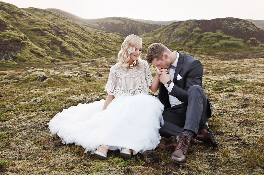 Bride and groomt seated on mossy landscape. Groom kisses bride's hand, evoking romance. Green hills in background.