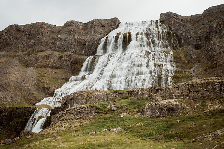 iconic iceland wedding location: Waterfall cascading down rocky cliffs with a person standing at the base. Lush green foreground, overcast sky.