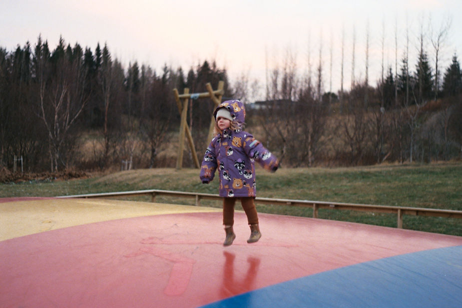 Child in a purple coat jumps on a colorful inflatable surface. Bare trees and grass in the background. Overcast sky, playful mood in iceland.