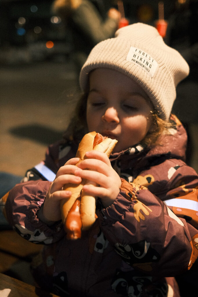 Child in winter coat and beanie joyfully holds hotdog, eyes closed in delight. Background is blurred, setting is outdoor iceland evening.