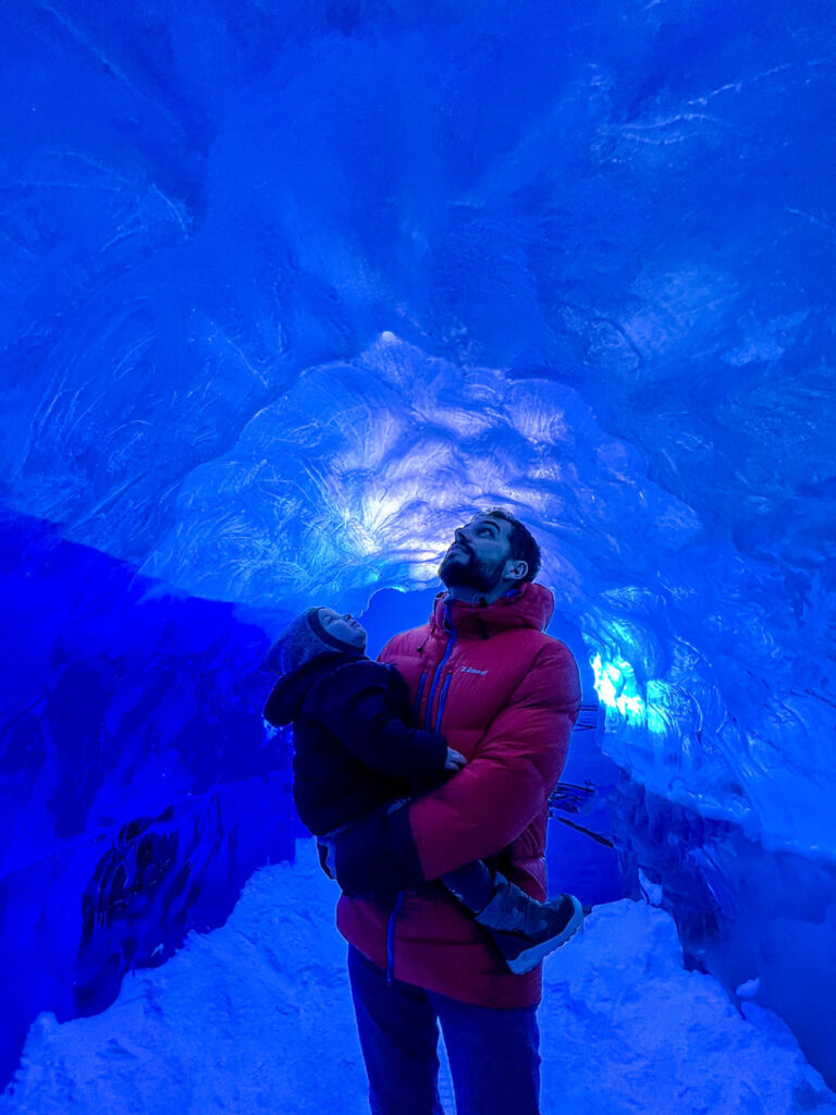 Man in red jacket holds a child inside a glowing blue ice cave in iceland, looking up. Cold, serene ambiance with rippling ice formations.