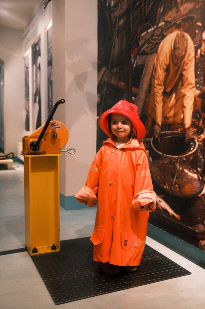 Young child in an orange raincoat and hat smiling indoors beside a yellow lever. Background features a vintage-style photo mural.