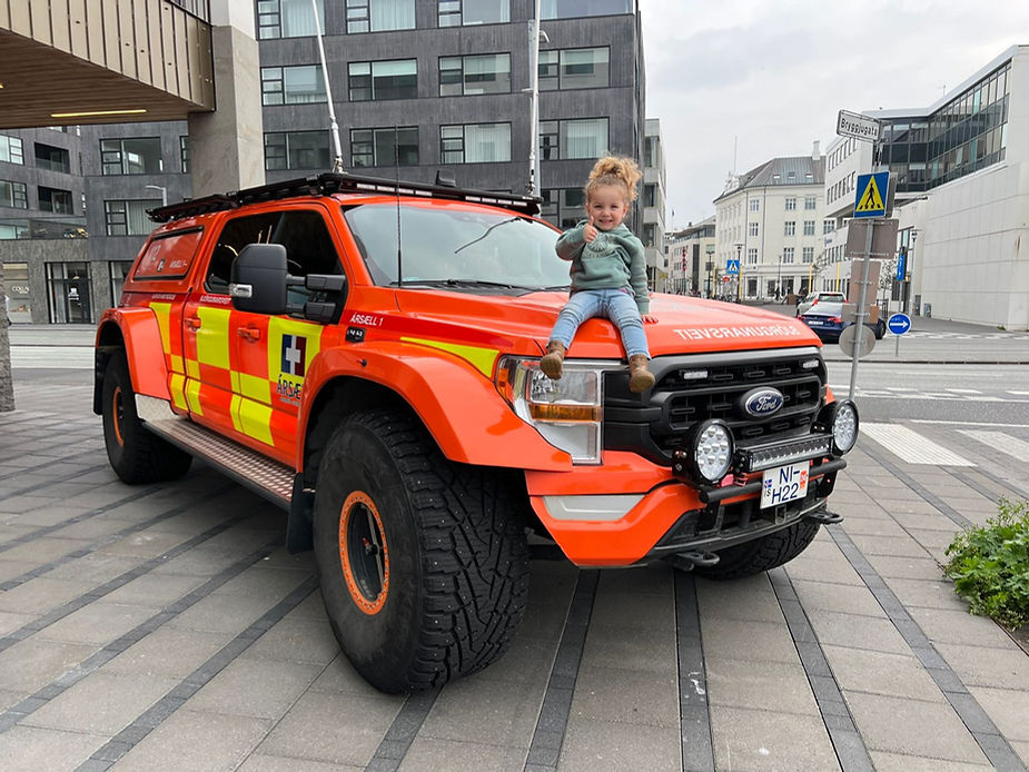 Child smiling on the hood of an orange rescue truck with yellow stripes, parked on a city street in iceland with modern buildings.