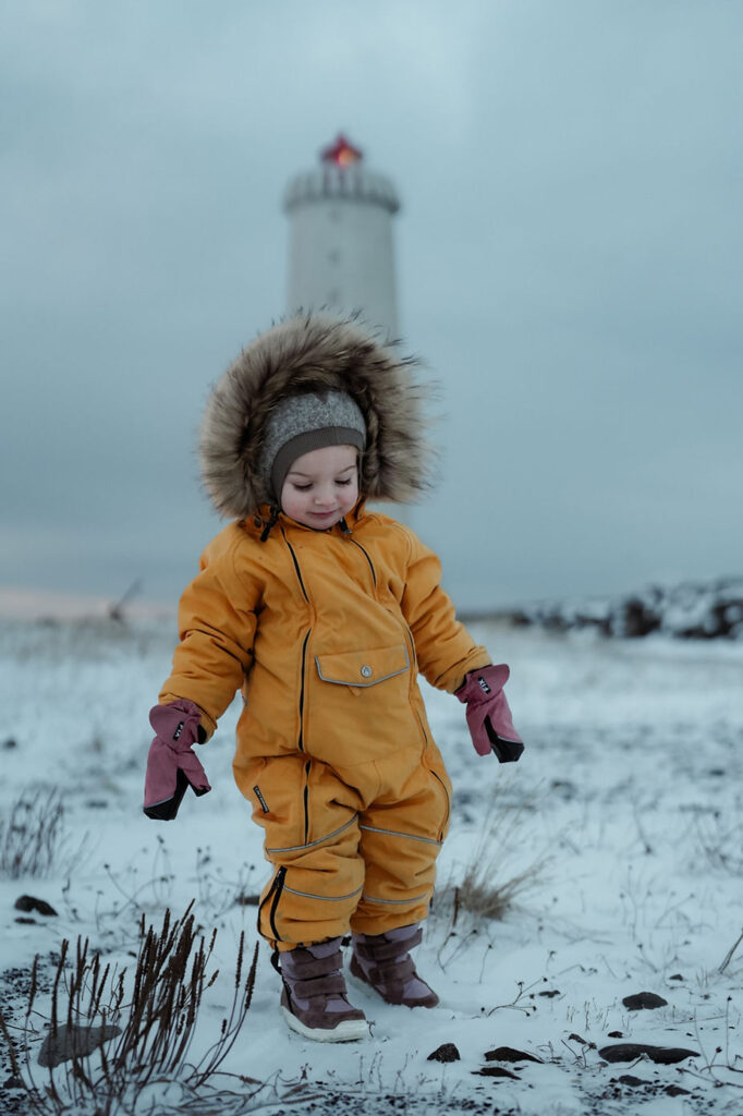 Child in a yellow snowsuit with fur hood and pink mittens stands in snowy field, lighthouse in background. Overcast skies, peaceful mood.