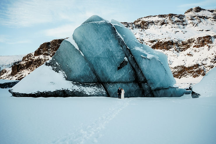 iconic iceland wedding location: Couple in wedding attire stands by a large ice formation in a snowy, mountainous landscape.
