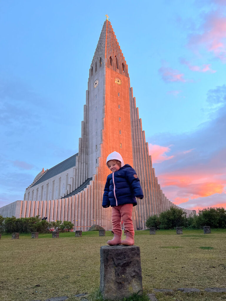 Child in a navy coat and pink pants stands on a stone post before a tall, sunlit iceland church at sunset, with a blue and pink sky backdrop.