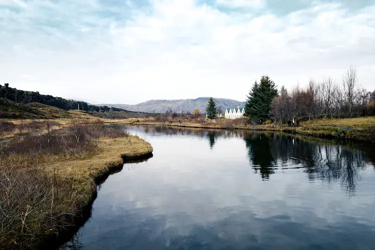 Tranquil landscape with a reflective pond, grassy banks, and snowy mountains in the background. Iceland wedding venue and trees line the right side.