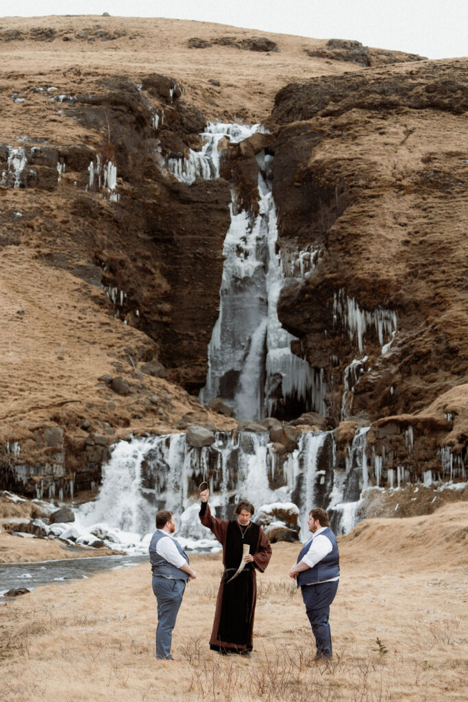 two grooms and officiant in front of an icy waterfall during Iceland Ásatrú elopement ceremony