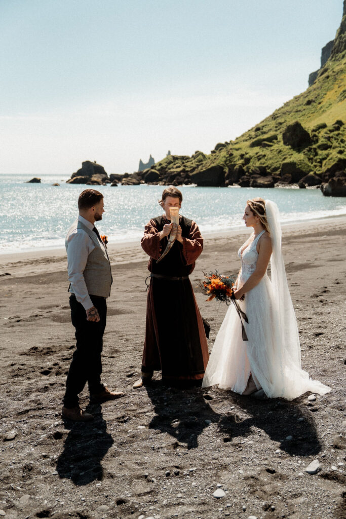 bride and groom on a sunny beach with rocky cliffs and blue sea. The bride holds flowers; the officiant, in robes, reads vows during Ásatrú elopement ceremony 
