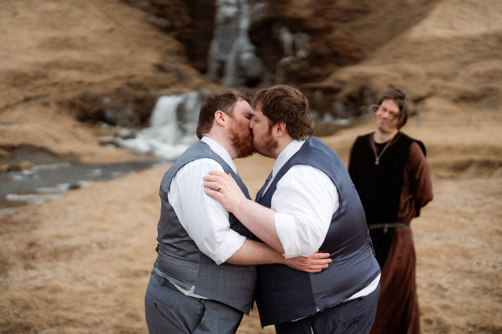 Two grooms in blue vests kiss outdoors, with a waterfall in the background. An officiant wearing a brown robe smiles nearby during Ásatrú elopement ceremony 