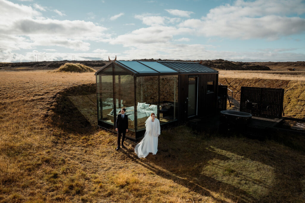 Bride and groom stand by a glass cabin in a vast, sunlit field under a blue sky before Ásatrú elopement ceremony 