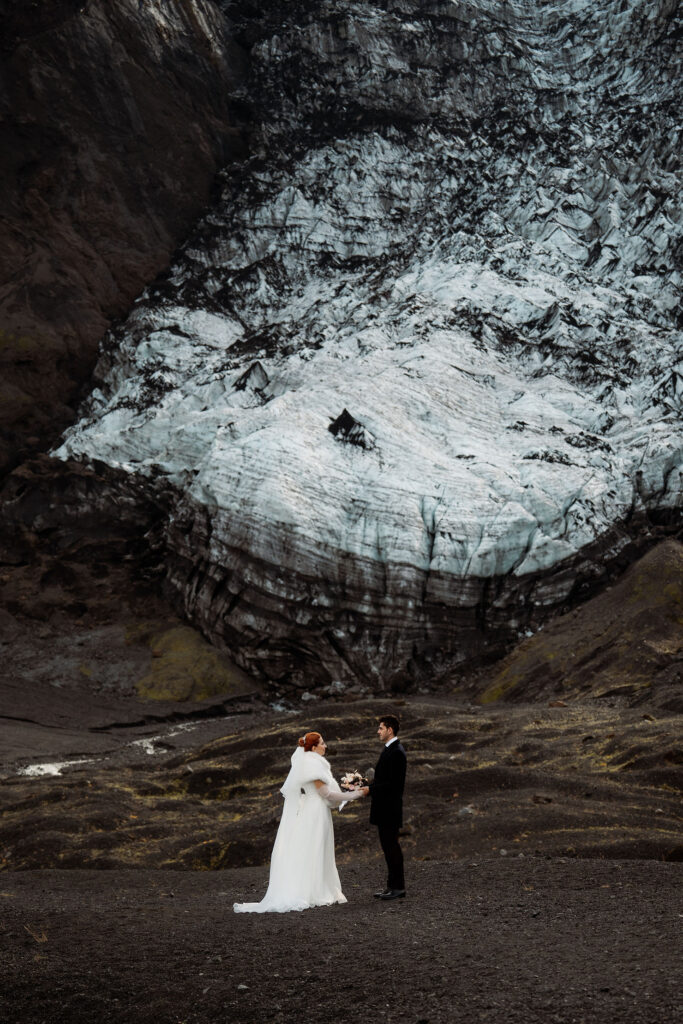 Bride and groom stand facing each other, holding hands, in front of a large glacier. Rugged terrain, moody setting for their Ásatrú elopement ceremony 