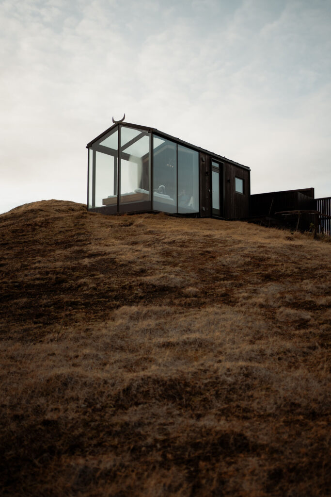 Modern glass cabin on a grassy hill under a cloudy sky. Minimalist design with a serene and tranquil atmosphere in iceland.