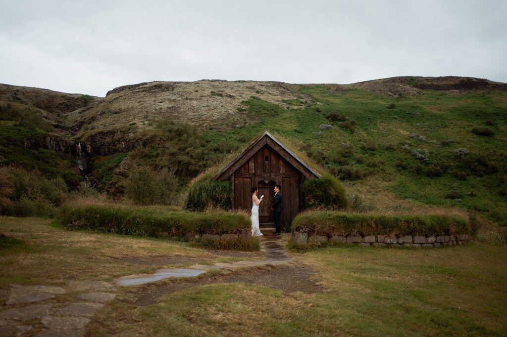 Bride and groom stand in front of a turf house. Green hills and overcast sky set a serene backdrop for the Ásatrú elopement ceremony.