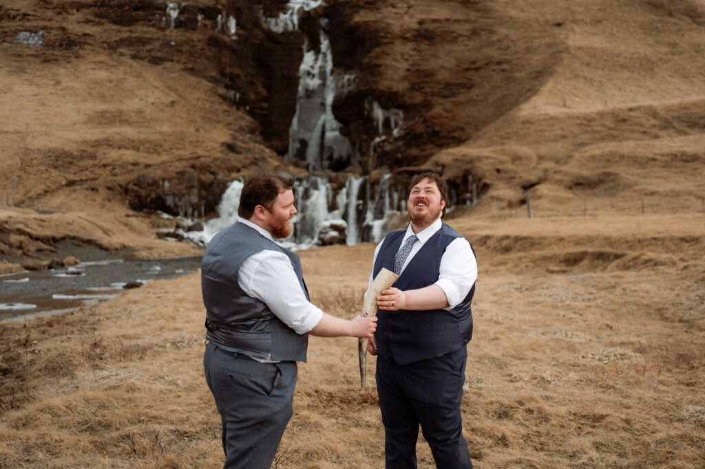 Two grooms smile joyfully, exchanging a horn against a backdrop of a waterfall and grassy hills. during Ásatrú elopement ceremony in iceland