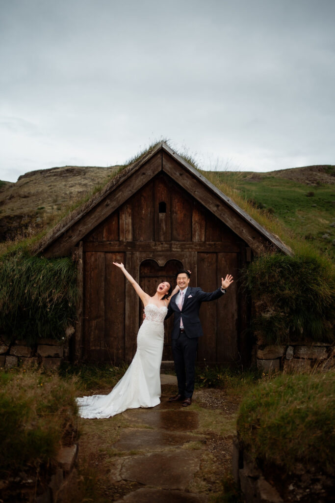 Bride and groom joyfully pose in front of a turf house with grassy roof amidst green hills for their Ásatrú elopement ceremony.