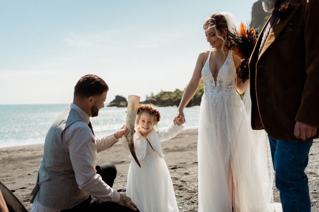 Bride holding child's hand on a sunny beach. Child with flower crown smiles, holding ornate horn during Ásatrú elopement ceremony