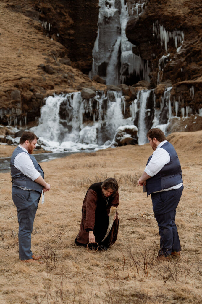 two grooms stand during elopement ceremony while officiant kneels, holding a horn and ring. 