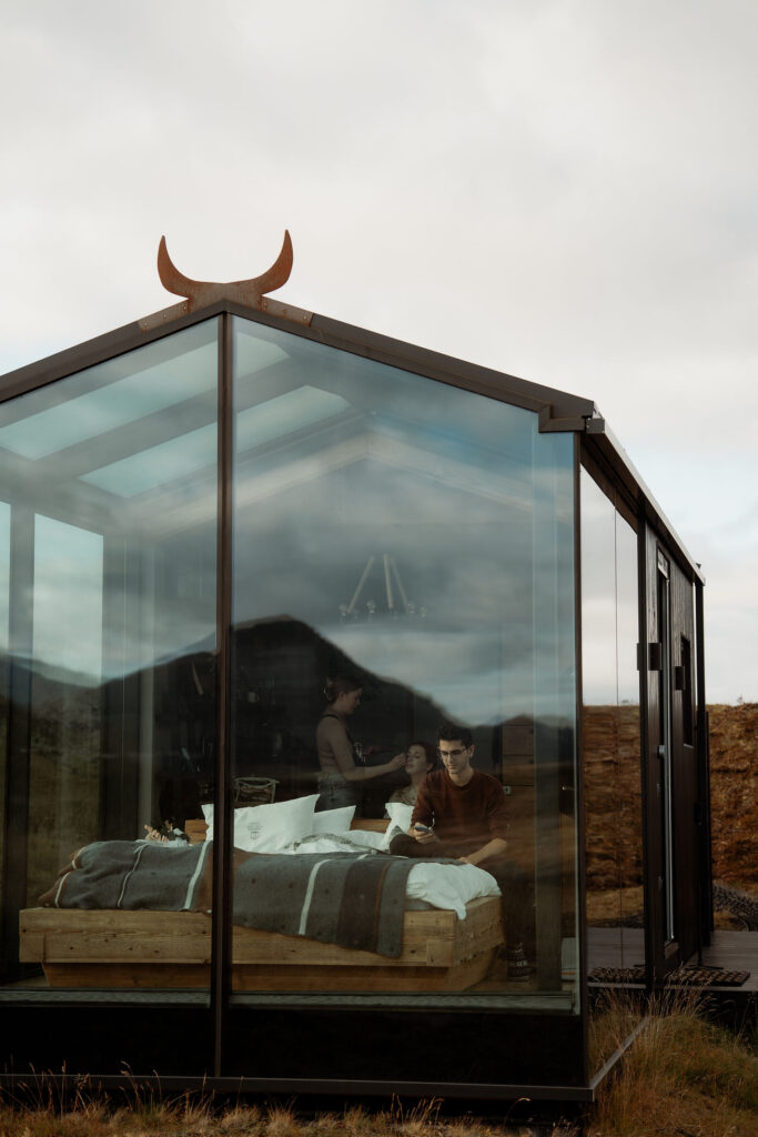 Transparent cabin; inside, a person sitting on a bed checks their phone, another gets makeup applied for Ásatrú elopement ceremony in iceland